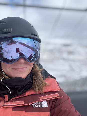 Heather in a black ski helmet and mirrored goggles reflecting a snowy mountain, smiling on a chairlift in a maroon North Face jacket, the real face behind the packing list